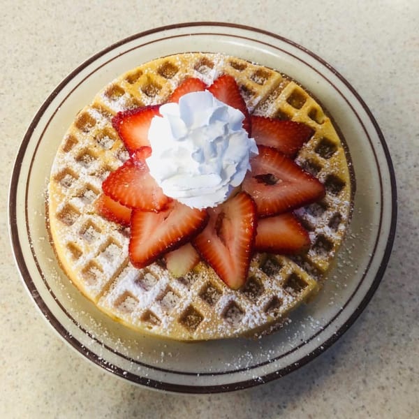 A golden waffle dusted with powdered sugar, topped with sliced strawberries and whipped cream.